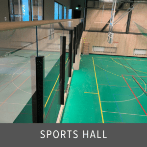 Frameless glass balustrade with dark posts and wooden top rail overlooking an indoor sports hall with green flooring and marked courts.