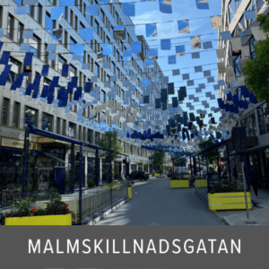Frameless glass panels used as partitions along a pedestrian street with cafés, restaurants and overhead art installation in central Stockholm.