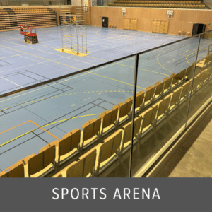 Clear frameless glass balustrade separating spectator seating from a blue indoor sports court with wooden bleachers and basketball hoops.