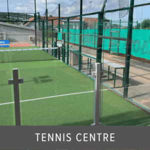 Glass fence panels with metal posts surrounding an outdoor tennis court with green synthetic turf, spectator area and windbreak netting.