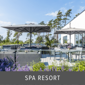 Frameless glass balustrade enclosing a sunny spa resort terrace with parasols, outdoor seating, dining tables and surrounding flower beds.