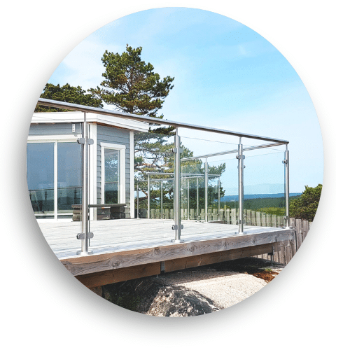 Stainless steel glass balustrade installed on a timber deck, with glass panels secured by clamps. A round handrail frames the terrace of a modern building, with a sea view and pine trees in the background.