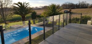 Modern glass pool fence with laminated clear glass and black aluminium posts surrounding a palm-lined pool area with open landscape views.