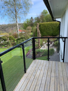Balcony with black aluminium and clear glass railing overlooking manicured garden and seating area in summer sun.