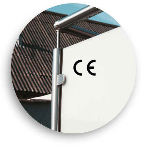 Close-up of CE-marked windbreak with opal glass and stainless steel posts, round handrail and clamp fixings, set against sky and tiled roof.