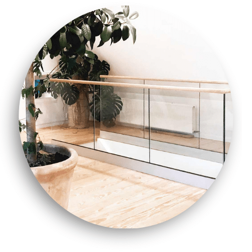 Frameless indoor glass balustrade with timber handrail, mounted on light wood flooring in bright open space with plants and daylight