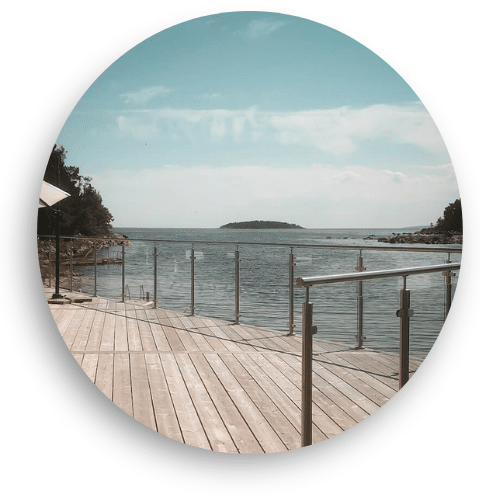 Glass balustrade with stainless steel posts installed along a wooden pier by the water. The railing ensures safety while maintaining open views of the sea and horizon.