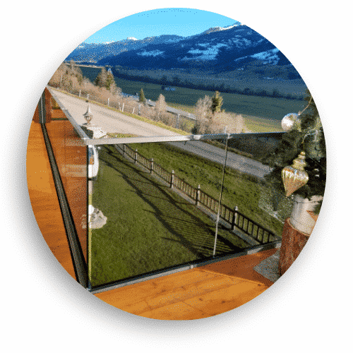 Modernt helglasräcke utan stolpar installerat på altan i trä, med fri sikt över ett lantligt landskap och snötäckta berg i bakgrunden.