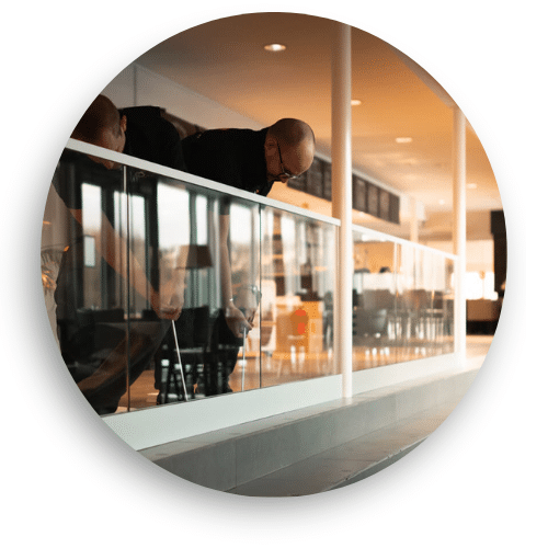 Two installers fitting a glass railing with white handrail and clamps in a bright public space, near a café with outdoor tables and chairs.