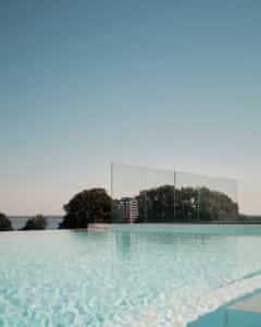 Premium frameless glass pool fencing around infinity pool with mirror-like water surface and unobstructed forest and ocean views.