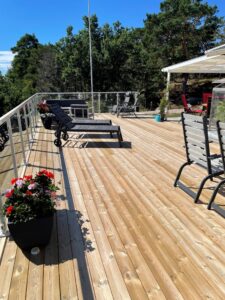 Large timber terrace with white aluminium and clear glass railing, furnished with loungers and garden furniture in sunlight.