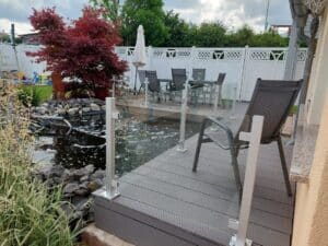 Sleek glass balustrade with white aluminium posts surrounding a small pond and outdoor seating area with red maple trees and classic white fencing.