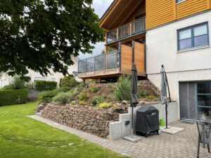 Raised decking with timber and black metal glass balustrade above stone retaining wall and landscaped sloping garden.