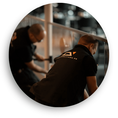 Two installers from Balustradedesign fitting a glass balustrade. Wearing black workwear with company branding, they fine-tune the fixings for secure installation. The glass reflects the interior surroundings.