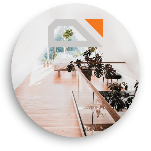 Glass railing with wooden elements along a loft, installed on timber flooring. Large windows and potted plants enhance the bright, natural atmosphere.