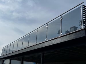 Modern garden balcony with smoked glass and black aluminium railing, surrounded by stone paving and trimmed hedges.