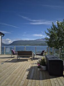 Spacious timber deck with glass railing and wooden posts, offering panoramic lake views and modern outdoor seating.
