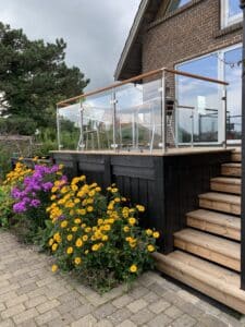 Deck balustrade in stainless steel and glass with timber handrail, floral foreground and black timber panel backdrop.
