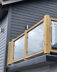 Glass balustrade with pressure-treated timber posts on corner balcony with grey façade and views of open sky.