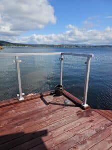 White aluminium and clear glass balustrade on red timber deck next to dock, with views of the sea and shoreline.
