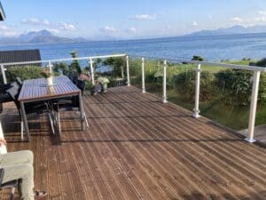 Spacious timber terrace with white aluminium glass railing and dining set, overlooking the ocean in sunny weather.