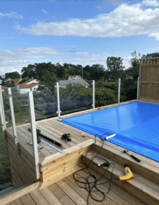 Glass balustrade under installation around elevated pool with white aluminium posts, wooden decking and bright blue pool cover.