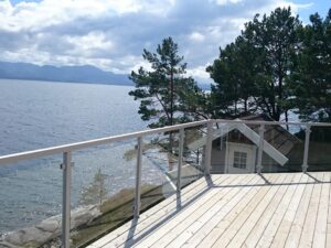 Aluminium and clear glass railing on light timber deck by the sea, facing open water and rocky coastal backdrop.