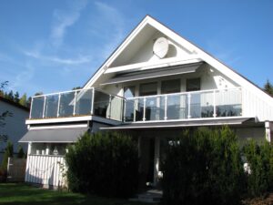 Balcony with white aluminium glass balustrade on pitched-roof villa, surrounded by greenery in the front garden.