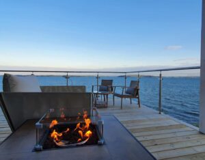 Stainless steel glass balustrade on coastal deck with lounge set and fire pit, overlooking water at golden hour.