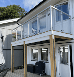 Glass railing with square white aluminium posts on wooden-decked balcony, attached to natural wood house façade.