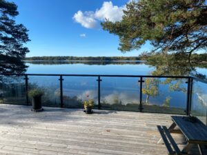 Black aluminium and clear glass balustrade framing lakefront view on timber terrace surrounded by forest and calm water.