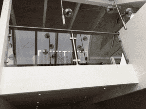 Glasräcke med rökfärgat glas och rostfri överliggare monterat på en vit loftkant. Stålfästen håller glaset på plats, och moderna ljuskällor reflekteras i glaset.