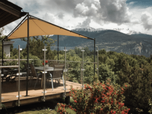 Adjustable wheeled deck with stainless steel glass balustrade and shade sail, overlooking a green valley and snowy peaks.