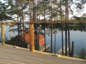 Timber deck with clear glass and wooden post balustrade by a lake, framed by pine trees and a red boathouse and jetty.