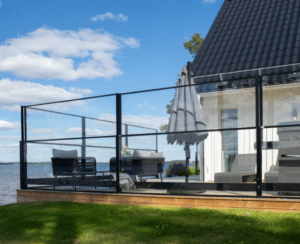 Outdoor space with black aluminium glass wind screen on timber deck near white house, with lake and cloud-filled blue sky.