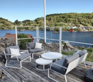 Cliffside terrace with clear glass and white aluminium railing, grey garden furniture and views of fjord, red cabins and hills.