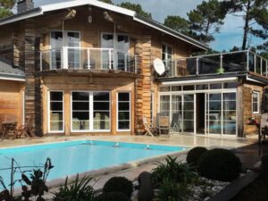 Tinted glass balustrade with stainless steel posts around log home’s balcony and pool, framed by pine trees and blue sky.